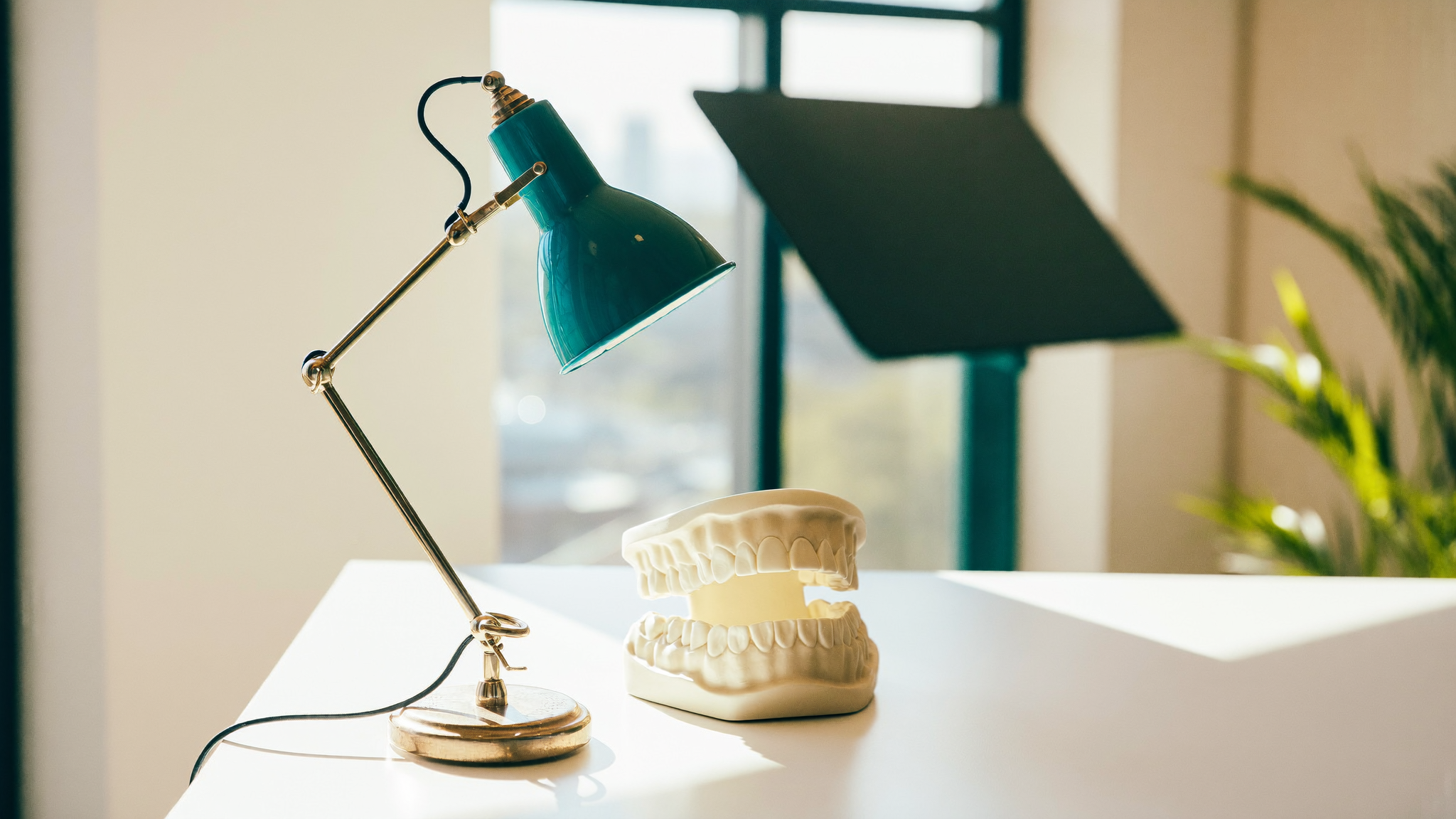 A dental study model of the upper and lower jaw on a bright planning desk beside a teal desk lamp, used to explain whether a tooth can be saved or should be replaced